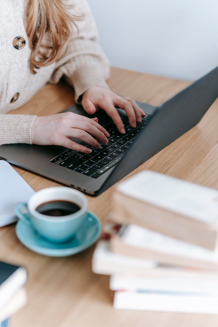 Crop anonymous female freelancer surfing netbook while sitting at wooden table with cup of hot coffee and books in room